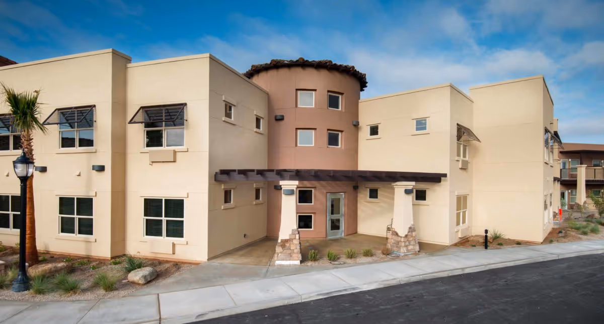 Exterior view of a modern two-story assisted living community building with beige and light brown walls, multiple windows, a small covered entrance supported by stone pillars, a palm tree, and a streetlamp along the sidewalk.