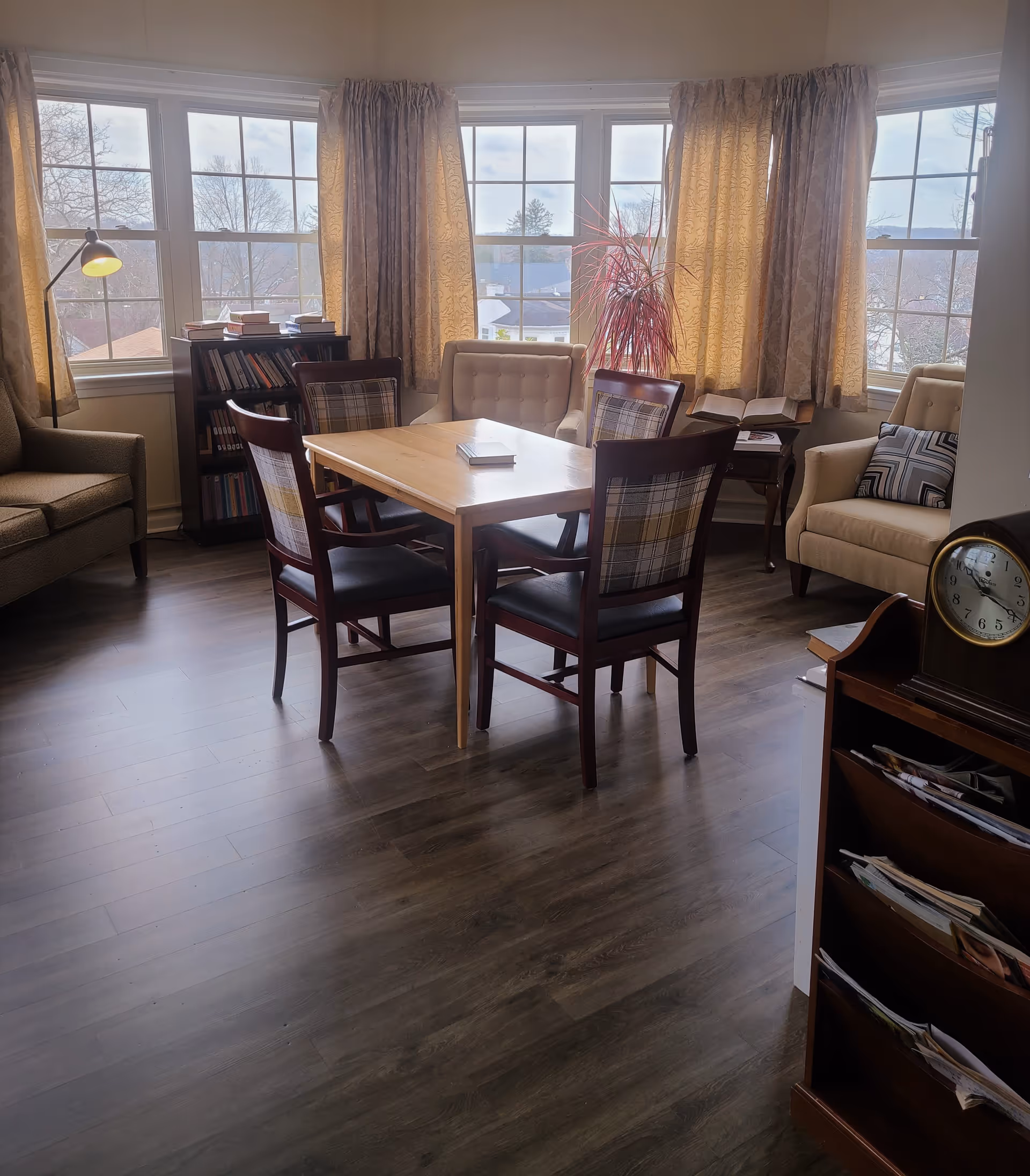 A cozy sitting area with a wooden table surrounded by four chairs with plaid backs, two beige armchairs with cushions, a small bookshelf filled with books, a floor lamp, and large windows with yellow curtains letting in natural light.