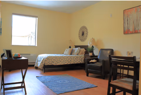 A well-lit bedroom with a bed covered in patterned bedding, a blue rug on the wooden floor, a dark armchair, a small table with a lamp, and a window letting in natural light. The walls are painted light yellow and decorated with a circular metal wall art and a framed painting.