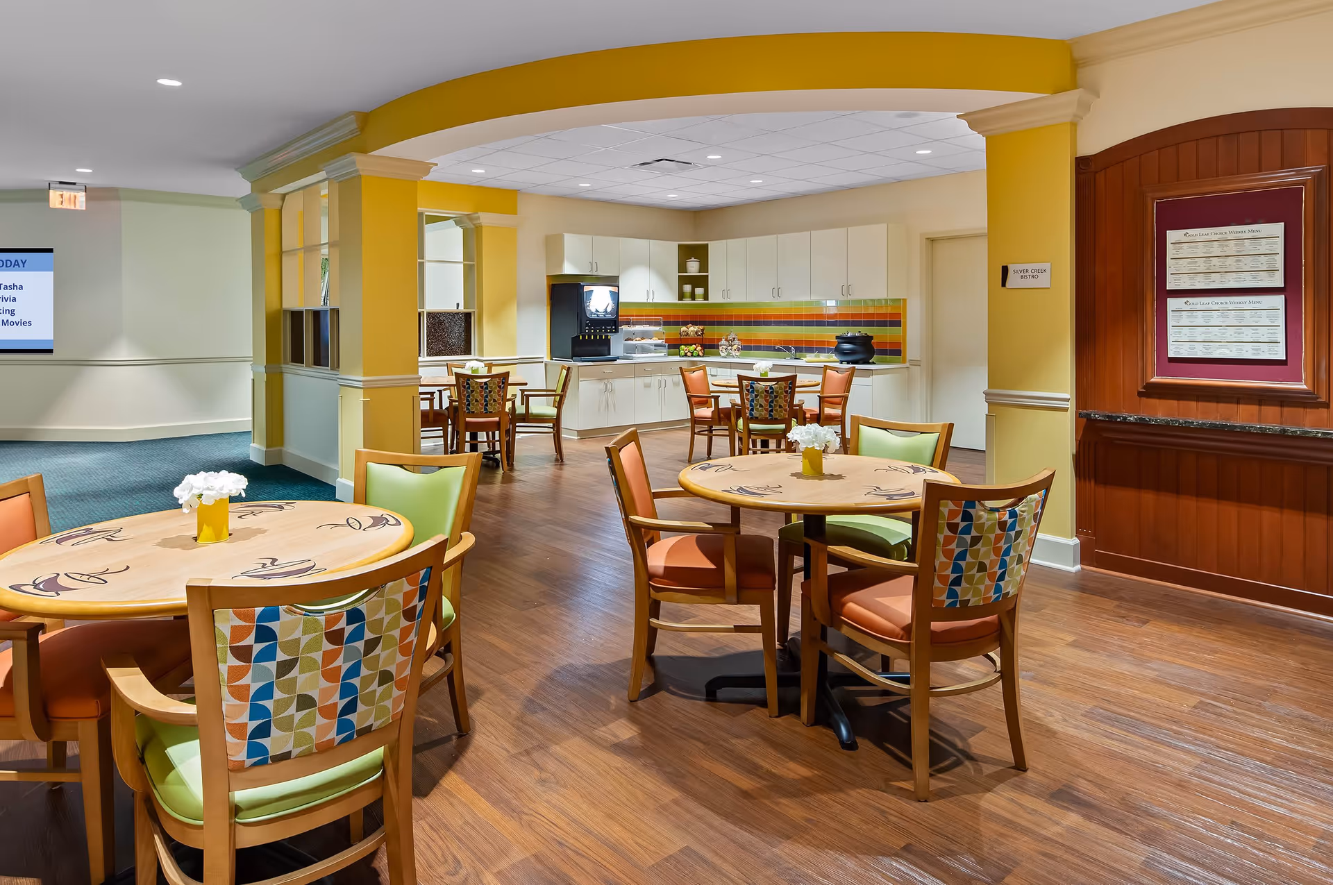 A bright and colorful dining area in a senior living facility with round wooden tables and chairs featuring multicolored patterned backs and green and orange cushions. The room has yellow walls with white trim, wood flooring, and a small kitchenette area with white cabinets and a colorful tiled backsplash. A sign on the wall reads 'Silver Creek Bistro' and a bulletin board displays menus.