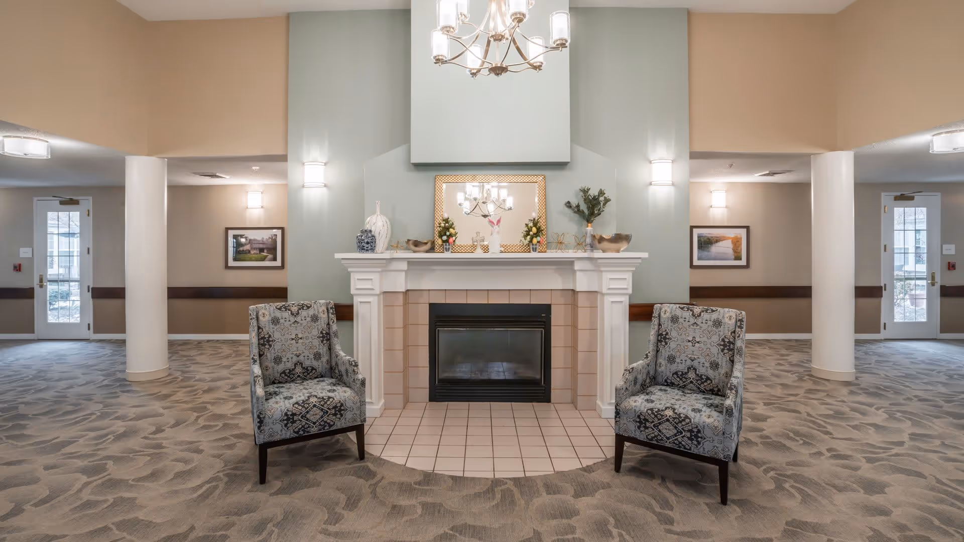A cozy seating area in a senior living facility featuring a white fireplace with decorative items on the mantel, including vases and plants. Two patterned armchairs are positioned symmetrically on either side of the fireplace. The room has soft lighting with wall sconces and a chandelier above, neutral-colored walls, and carpeted flooring with a subtle pattern. Doors and framed pictures are visible in the background.
