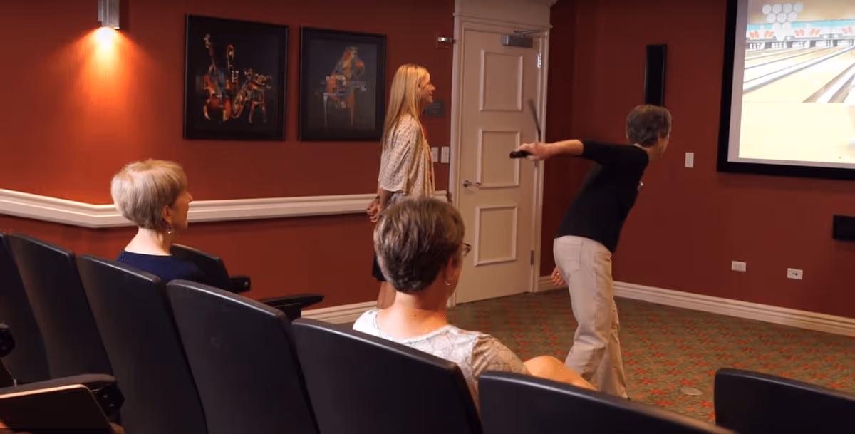 A group of elderly women in a room with red walls and carpeted floor, some seated in black chairs and one woman standing and bowling towards a large screen displaying a bowling game.