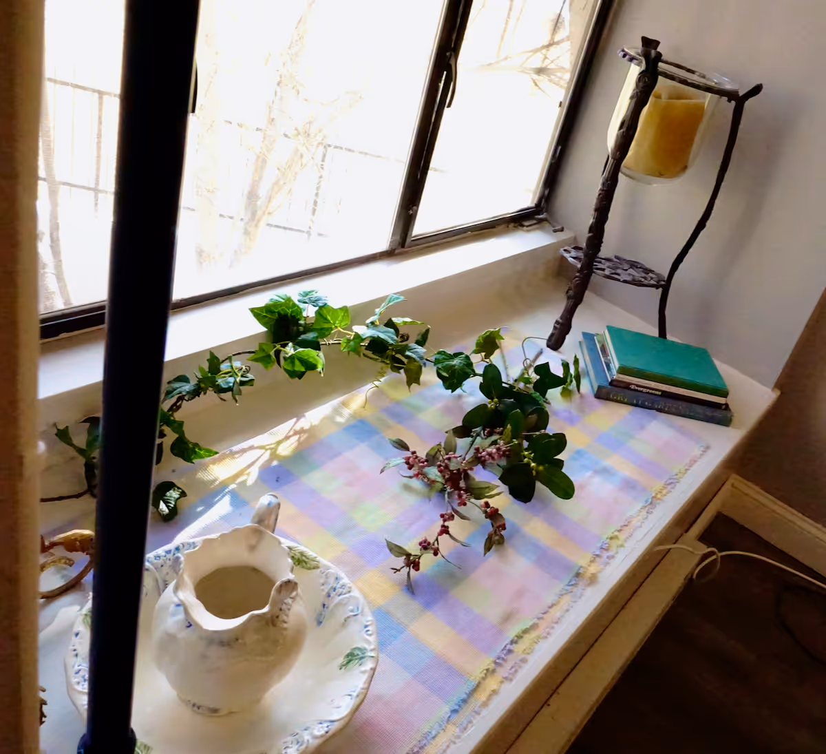 Sunlit windowsill vignette with a pastel checkered cloth, a ceramic pitcher and bowl, trailing greenery, stacked books, and a decorative candle holder.