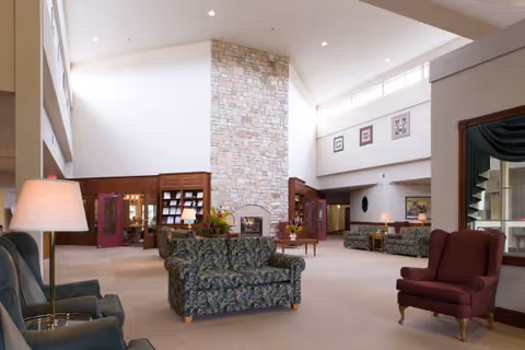 Spacious senior living facility common lounge with sofas and chairs arranged around a central stone fireplace under a high vaulted ceiling.