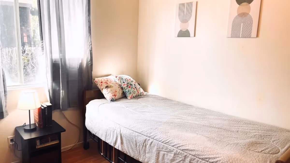 A small bedroom with a single bed covered with a light gray patterned blanket and two floral pillows. Next to the bed is a black nightstand with a lamp and some books. A window with sheer gray curtains allows natural light to enter the room. Two abstract art pieces hang on the beige wall above the bed.