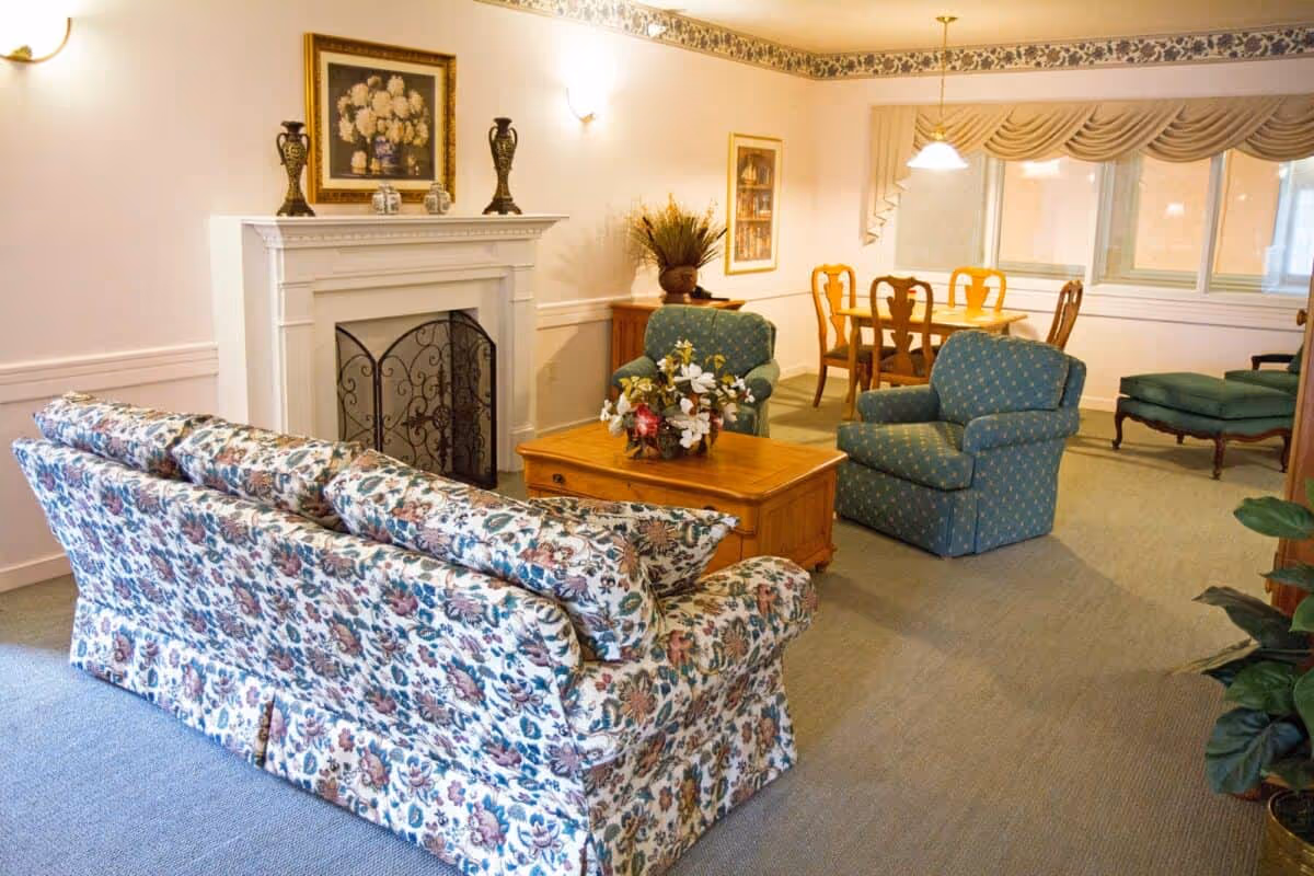 A cozy living room area featuring a floral-patterned sofa, two blue armchairs with a subtle pattern, a wooden coffee table with a floral centerpiece, and a white fireplace with decorative vases and a framed floral painting above it. In the background, there is a wooden dining table with six chairs under a hanging light fixture, and large windows with beige drapes.