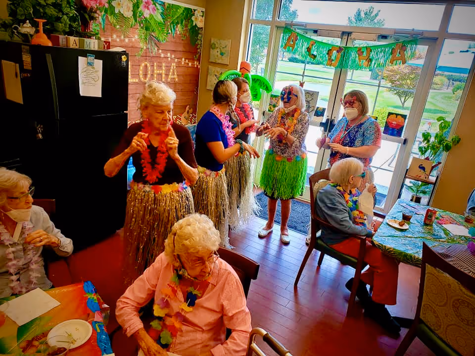 A group of elderly women dressed in colorful Hawaiian-themed attire, including grass skirts and leis, are gathered in a room decorated with tropical decorations and a banner that reads 'ALOHA'. Some women are seated at tables with food and drinks, while others stand and interact near a large window with a view of greenery outside.