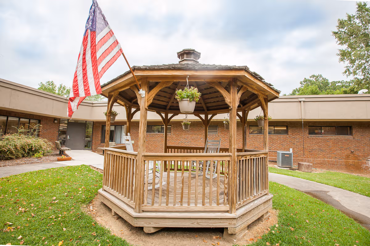 A wooden gazebo with hanging flower pots and rocking chairs inside, situated on a grassy area in front of a brick building. An American flag is mounted on the gazebo, and a paved walkway curves around it.
