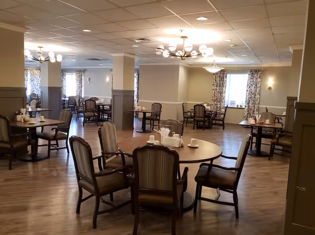 A spacious dining room with multiple round and square tables surrounded by cushioned chairs. The room has wooden flooring, beige walls with wainscoting, and several windows with patterned curtains allowing natural light. Modern light fixtures hang from the ceiling, and tables are set with cups, napkins, and condiments.