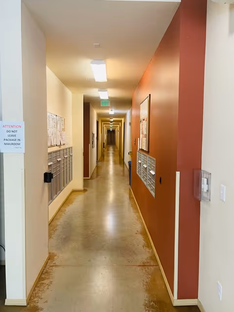 A long, narrow hallway in a senior living facility with mailboxes mounted on both walls. The floor is polished concrete, and the walls are painted white and reddish-brown. Fluorescent lights illuminate the corridor, and there are various notices and papers posted above the mailboxes.