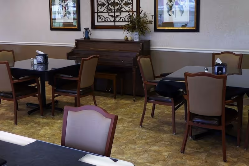 Interior view of a dining area with several tables and chairs. A wooden piano with a small bench is against the wall, decorated with a plant and framed artwork above it. The floor has a patterned linoleum surface.