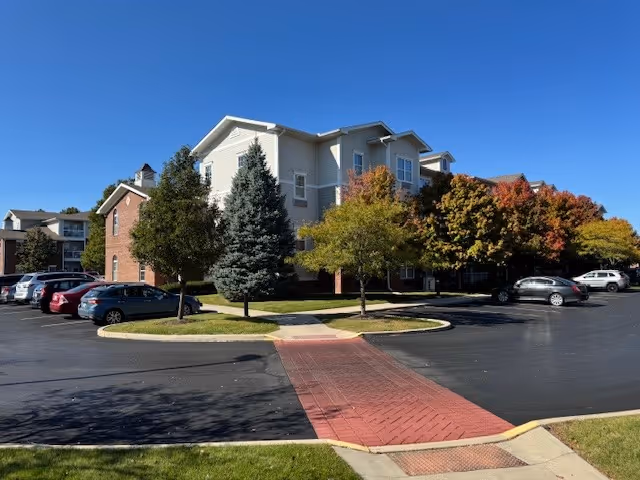 Front exterior of a multi-story senior living building with parked cars, trees, and a brick walkway under a clear blue sky.