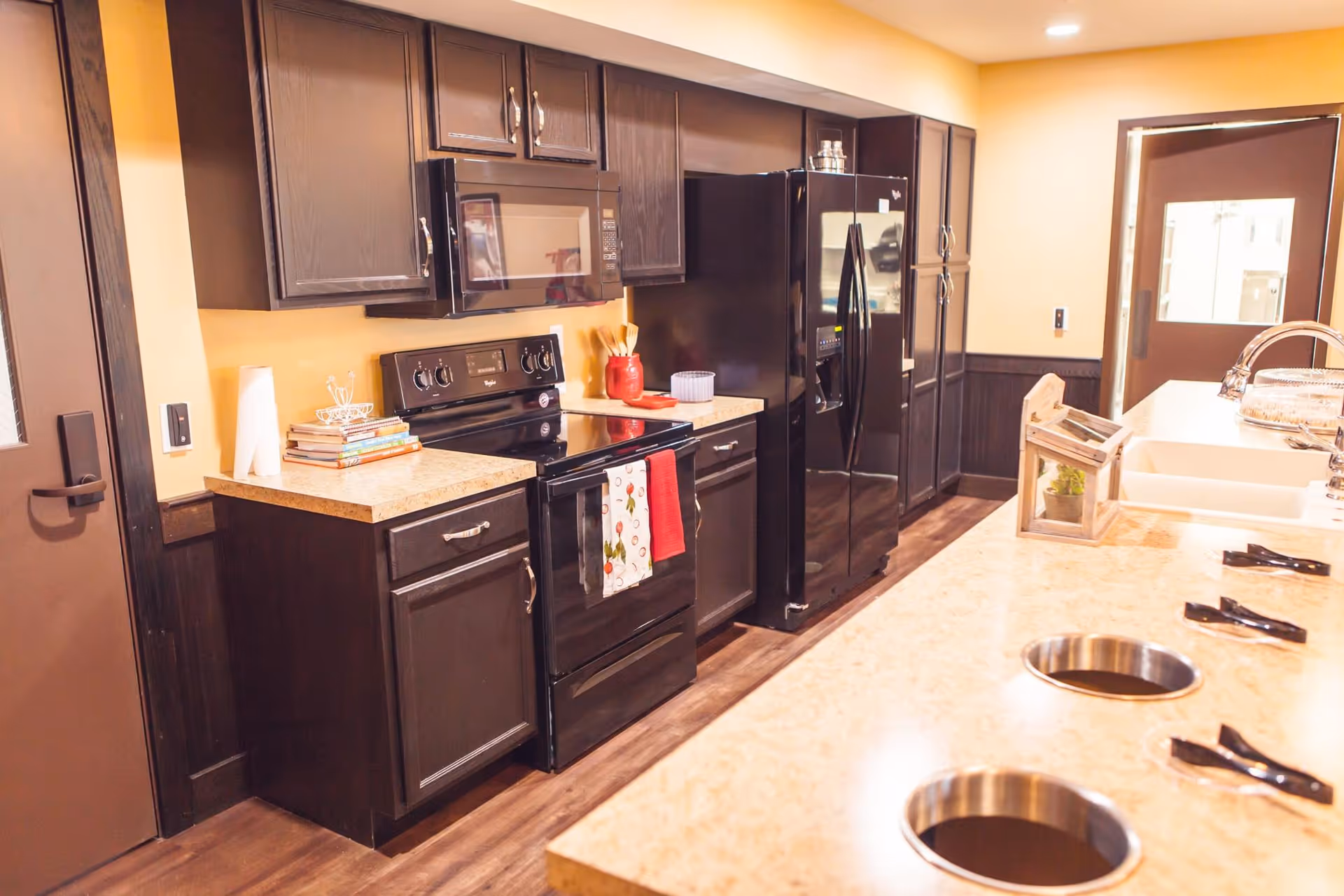 A modern kitchen with dark wood cabinets, a black stove and microwave, a black refrigerator, and a beige countertop island with built-in sinks and faucets. The kitchen has warm yellow walls and a wooden floor.