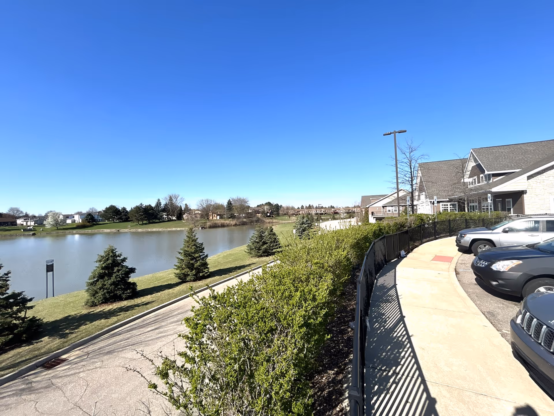 View of a lakeside area with a clear blue sky, several evergreen trees, and a calm body of water. On the right side, there is a paved sidewalk next to a black metal fence, parked cars, and part of a building with a sloped roof.