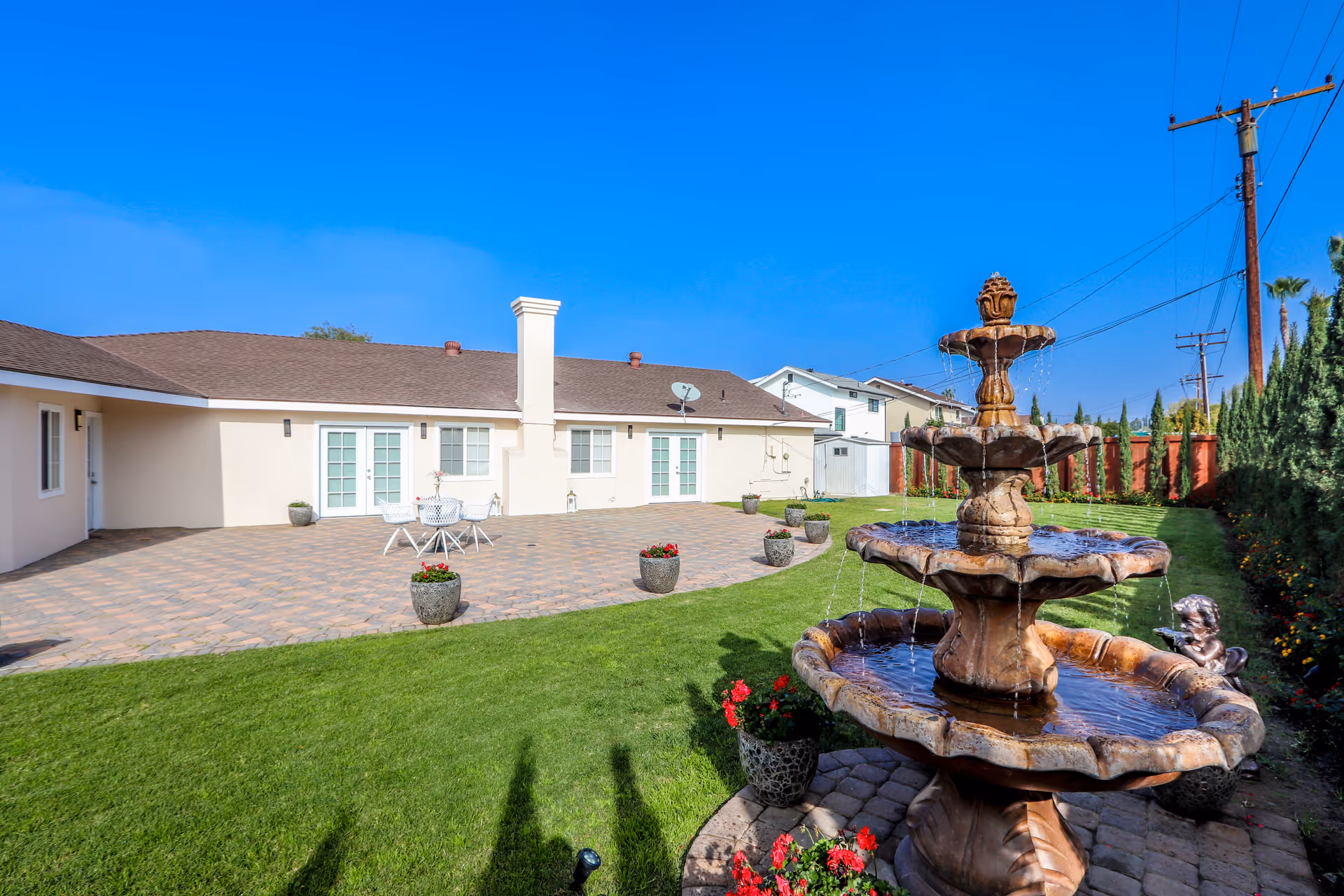 Outdoor view of a senior living facility showing a well-maintained lawn with a three-tiered water fountain in the foreground, potted plants with red flowers, a paved patio area with white outdoor furniture, and a beige building with multiple doors and windows under a clear blue sky.