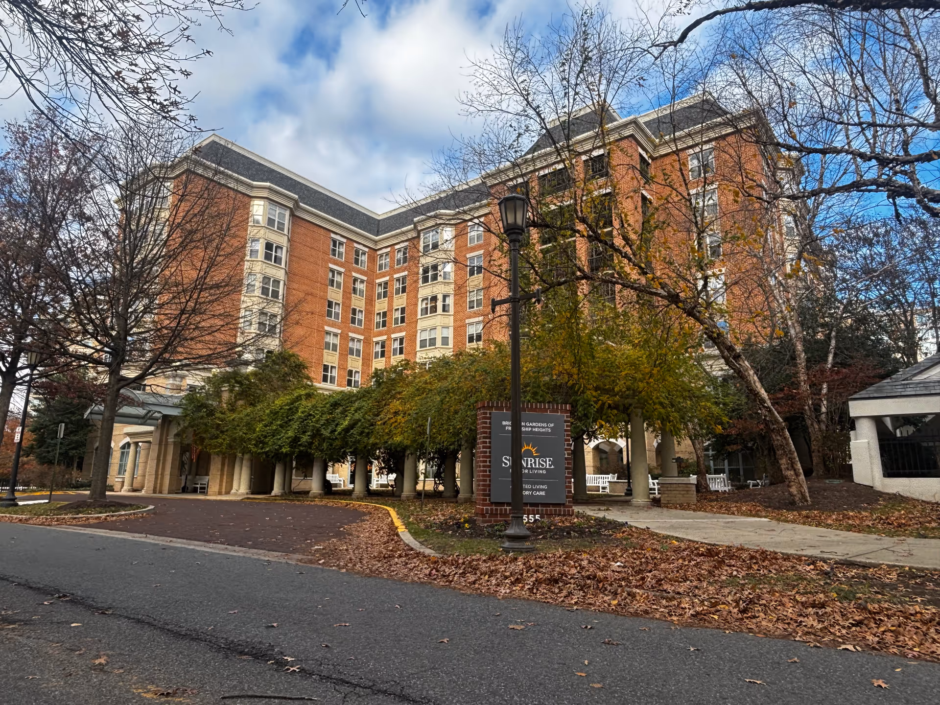 Exterior view of a multi-story brick building with white window frames and a covered entrance surrounded by trees with autumn leaves. A sign near the entrance reads 'Brighton Gardens of Friendship Heights Sunrise Senior Living Assisted Living Memory Care.' The sky is partly cloudy.