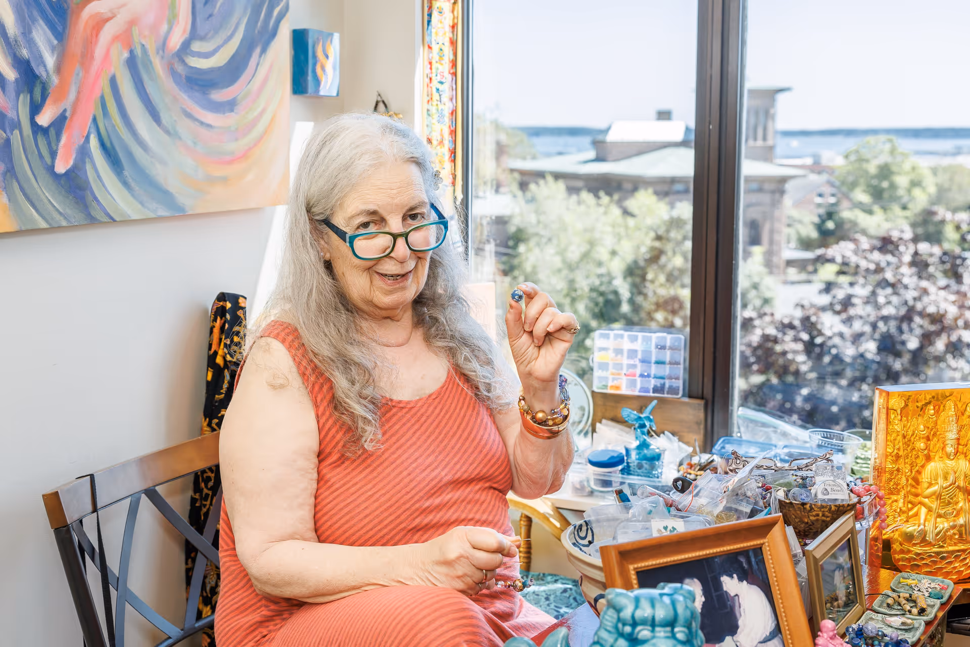 An elderly woman with long gray hair and teal glasses sits on a chair near a large window, holding a small bead or object in her hand. She is wearing a sleeveless orange dress and is surrounded by various decorative items, framed photos, and craft supplies on a table. Outside the window, trees and buildings are visible under a clear sky.