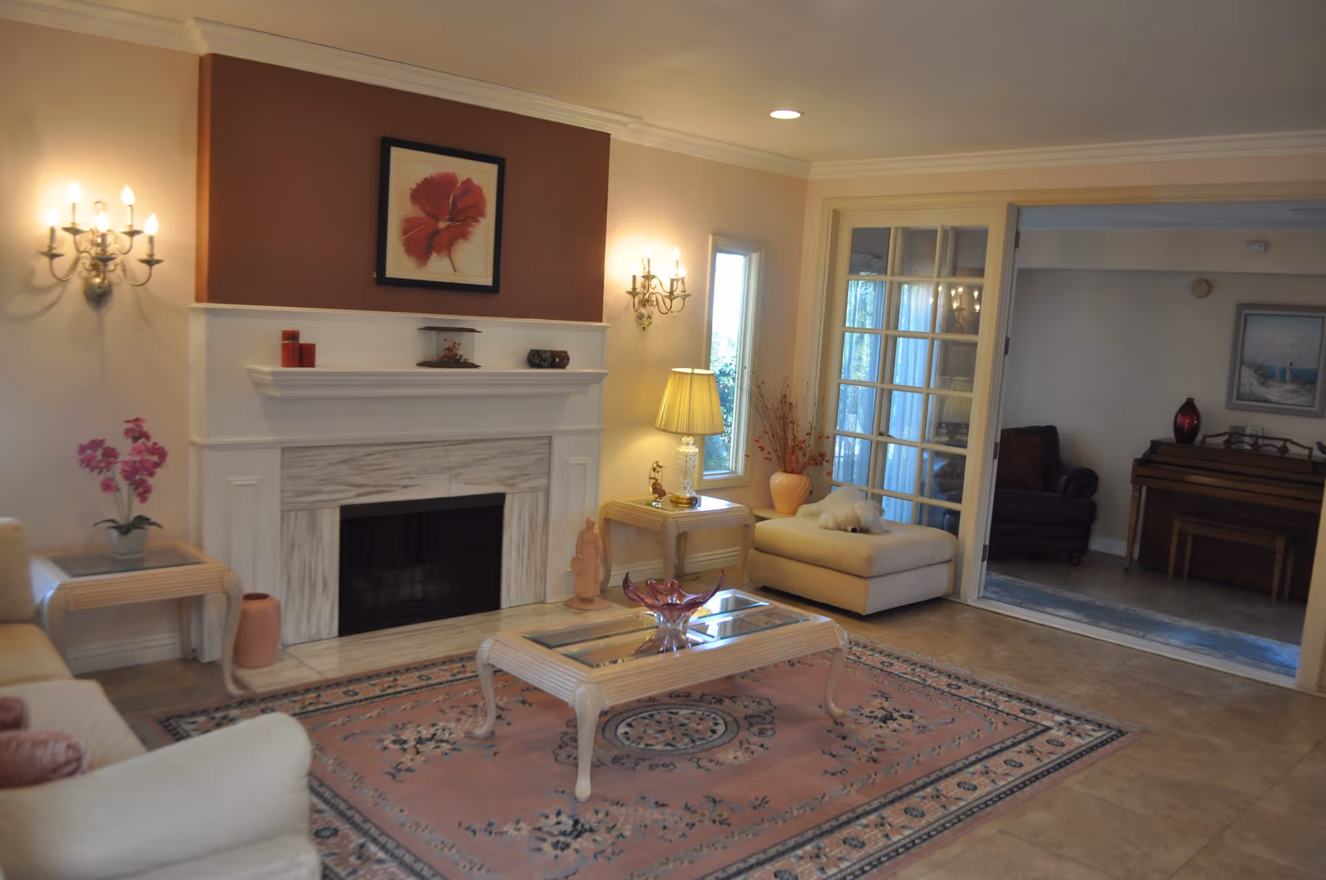 A cozy living room with a white fireplace centered on a wall painted in a warm brown and cream color scheme. The room features a pink patterned area rug, a glass-top coffee table, a beige ottoman with a white stuffed animal on it, and a side table with a lamp and decorative items. There are wall sconces on either side of the fireplace and a framed flower painting above it. In the background, a second room with a piano and armchair is visible through glass-paneled sliding doors.