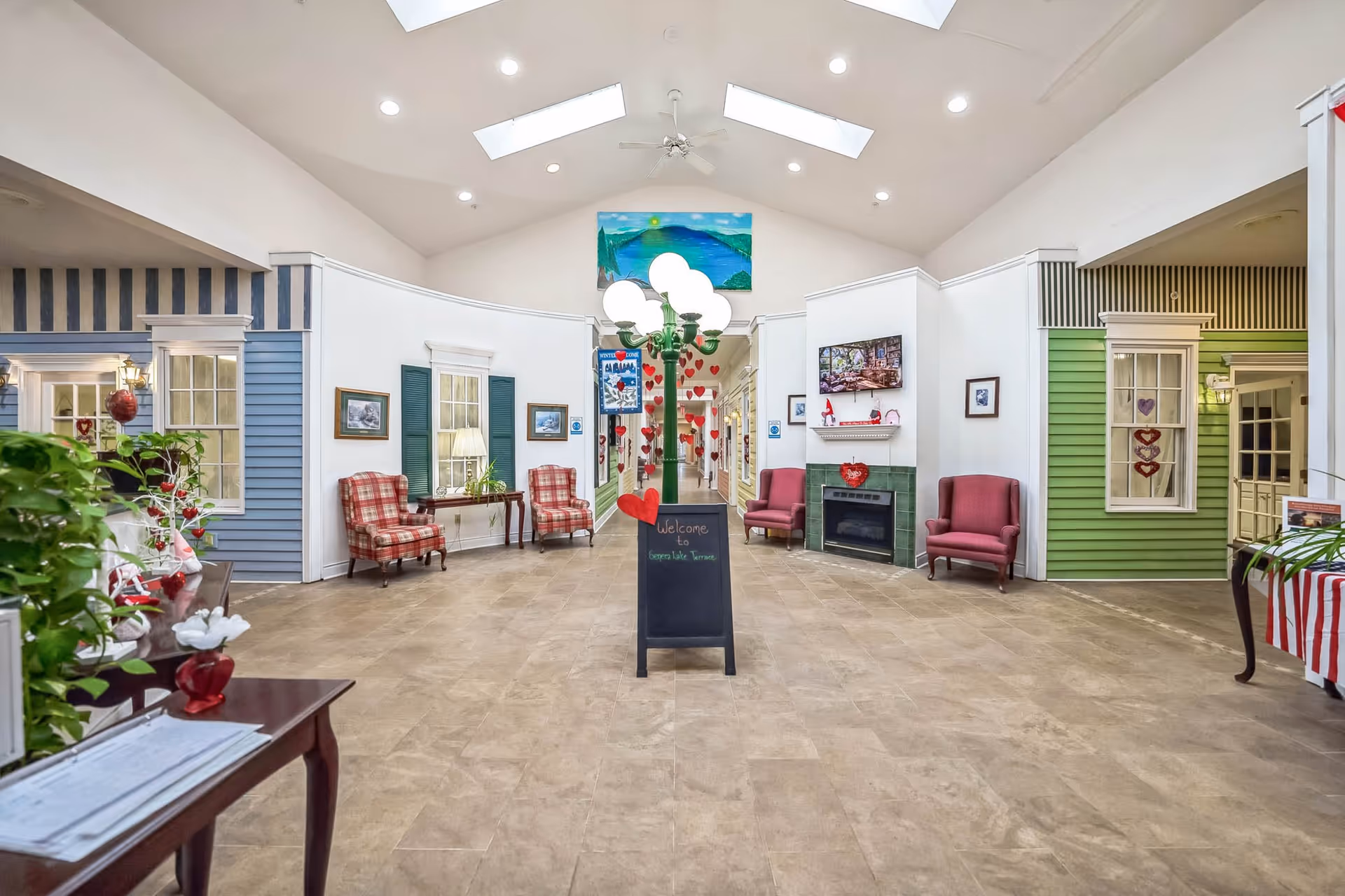 A bright and spacious interior hallway of Seneca Lake Terrace with a high ceiling and skylights. The hallway is decorated with red heart-shaped ornaments hanging from a central lamp post. There are cozy seating areas with red plaid and solid red armchairs, small tables with lamps, and framed pictures on the walls. The walls are painted in white with sections of blue and green siding, resembling a small village street. A blackboard sign in the center welcomes visitors to Seneca Lake Terrace.