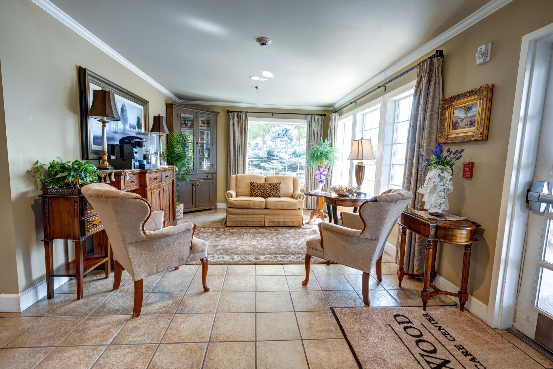 A bright and cozy sitting area in a senior living facility with two beige armchairs facing a small sofa. The room features large windows with curtains, a patterned rug, wooden furniture including a sideboard with lamps and plants, a corner cabinet, and a small table with a lamp and decorative items. A door with a mat labeled 'WOOD CARE CENTER' is visible on the right side.