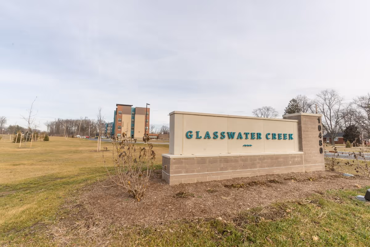 Outdoor view of the entrance sign for Glasswater Creek with the building visible in the background, surrounded by grass and some small trees under a cloudy sky.