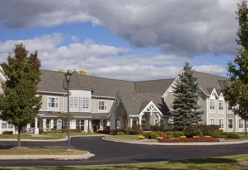 Front view of a two-story senior living building with a covered entrance, circular driveway, and landscaped grounds under a cloudy sky.