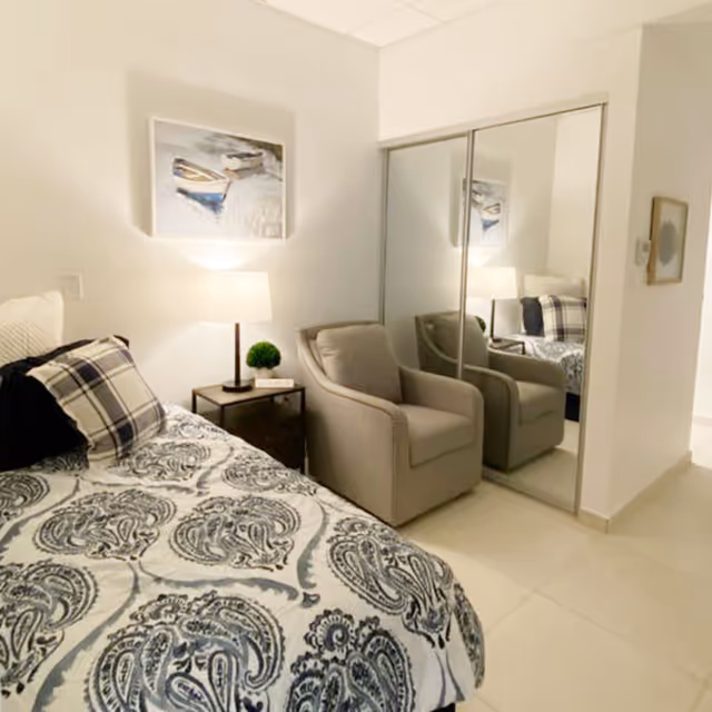 A cozy bedroom featuring a bed with a patterned bedspread and plaid pillows, a bedside table with a lamp and a small plant, a comfortable armchair, and a mirrored closet door reflecting the room's furnishings. A framed picture of boats hangs on the wall above the lamp.