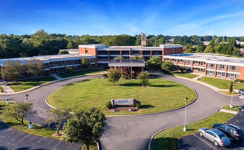 Aerial view of Altenheim Family-first Senior Living facility showing a large U-shaped brick building surrounding a circular driveway with a grassy center. Several cars are parked in the parking lot, and trees and greenery surround the property under a clear blue sky.