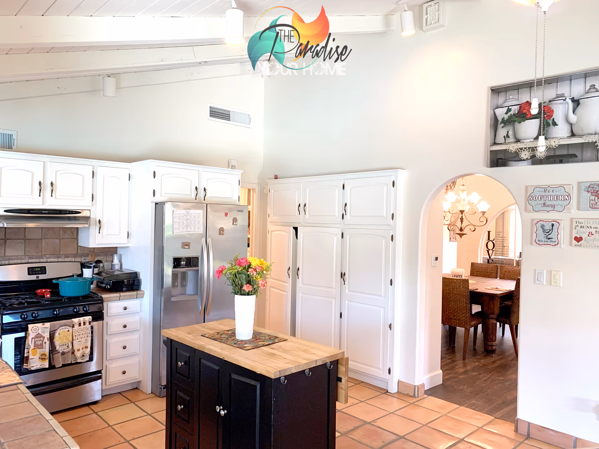 Bright kitchen with white cabinets, stainless steel refrigerator, small black island topped with a vase of flowers, and an arched entry to a dining room.