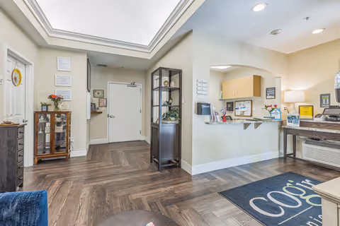 Interior view of a senior living facility reception area with a front door, a reception desk with flowers and informational materials, a display cabinet with decorative items, and wood-patterned flooring. The ceiling has recessed lighting and a skylight.