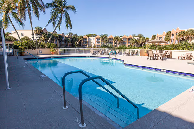 Outdoor swimming pool with metal handrails, lounge chairs and palm trees in front of apartment-style buildings.