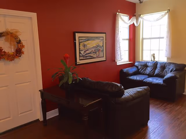 A cozy living room with two black leather sofas arranged around a dark wooden table with a potted plant. The walls are painted red and beige, with a framed painting on the red wall and two windows with white curtains letting in natural light. A door decorated with a fall-themed wreath is visible on the left.