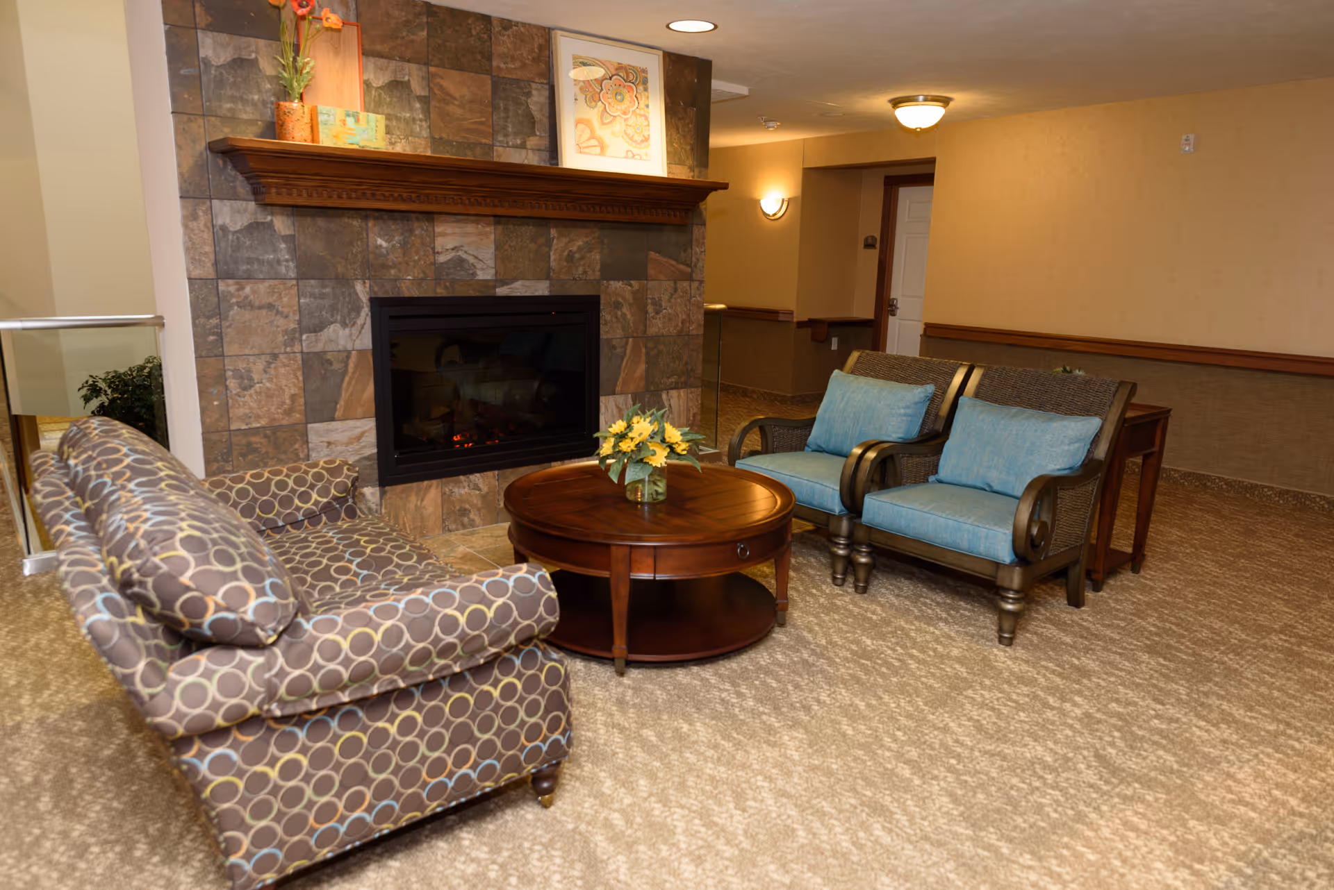 A cozy seating area in a retirement facility featuring a patterned armchair, two wooden chairs with blue cushions, a round wooden coffee table with a small flower arrangement, and a stone-tiled fireplace with a wooden mantle displaying decorative items and framed artwork.