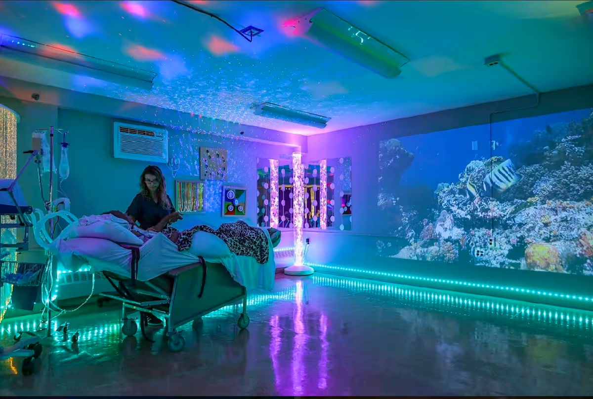 Sensory therapy room interior with a hospital bed and IV stand, colorful LED lighting, a wall projection of an underwater reef, and a caregiver seated beside the bed.