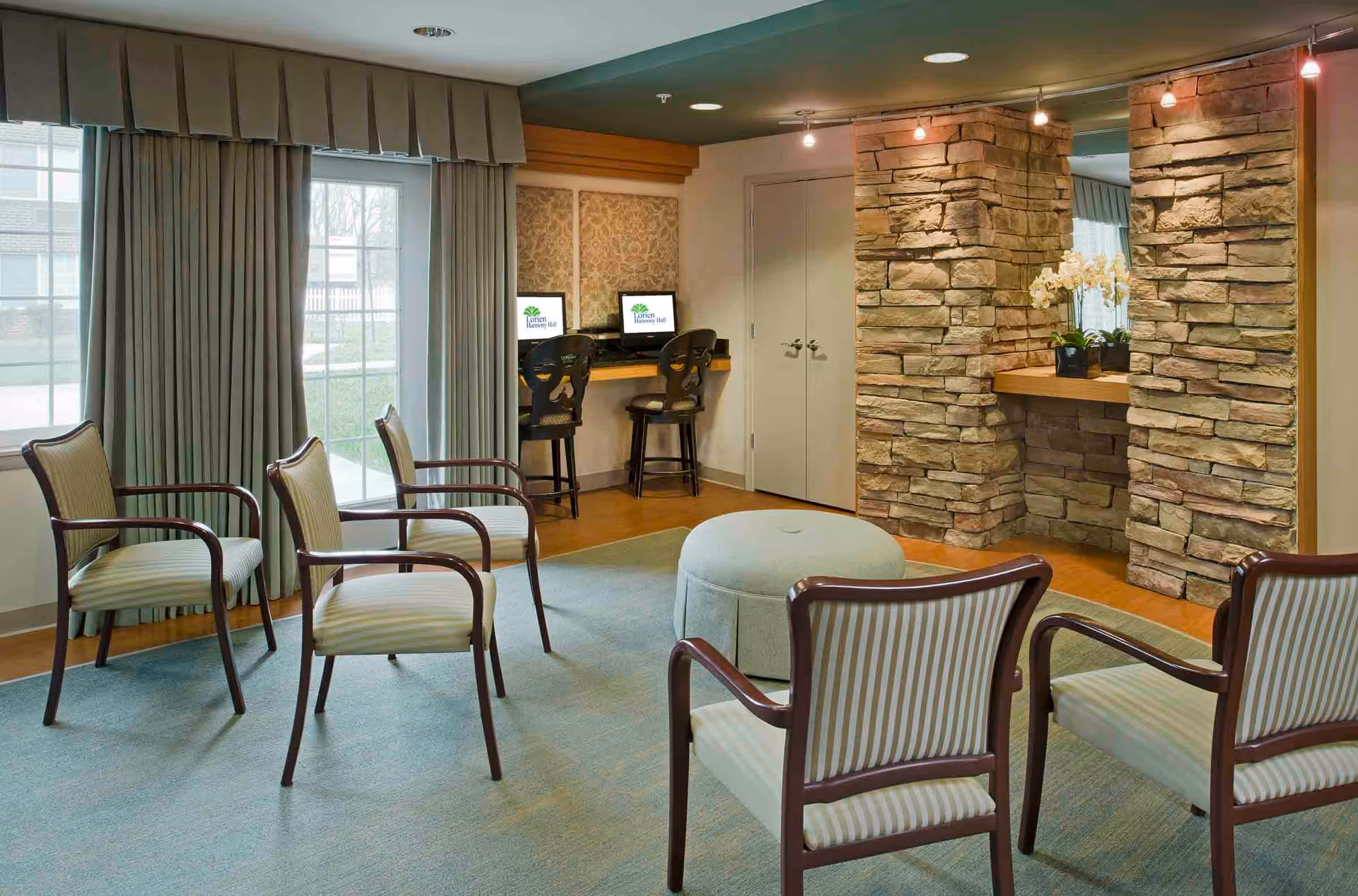 A cozy common area in Lorien Harmony Hall featuring six striped armchairs arranged around a central round ottoman on a light blue carpet. The room has large windows with curtains, a stone accent wall with a mirror and potted orchids, and a small computer workstation with two chairs and monitors displaying the Lorien Harmony Hall logo.