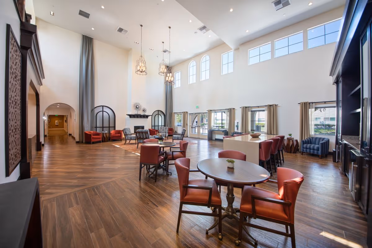 Spacious senior-living common room with high ceilings, multiple seating areas, tables, and large windows.