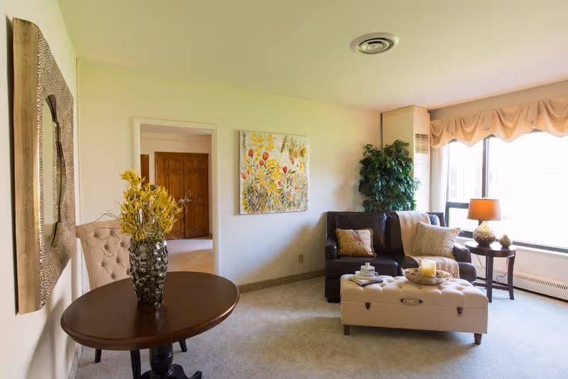 A cozy living room with a dark brown loveseat adorned with pillows and a throw blanket, a tufted beige ottoman serving as a coffee table with decorative items on top, a side table with a lamp, a large window with beige curtains allowing natural light, a potted plant in the corner, a round wooden table with a vase of yellow flowers, and a colorful floral painting on the wall.