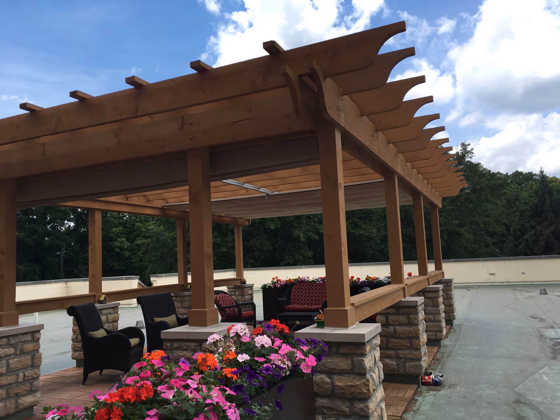 Outdoor seating area with a wooden pergola structure supported by stone pillars. There are several cushioned chairs and a sofa inside the pergola. Colorful flowers in planters are in the foreground, and trees and a partly cloudy sky are visible in the background.