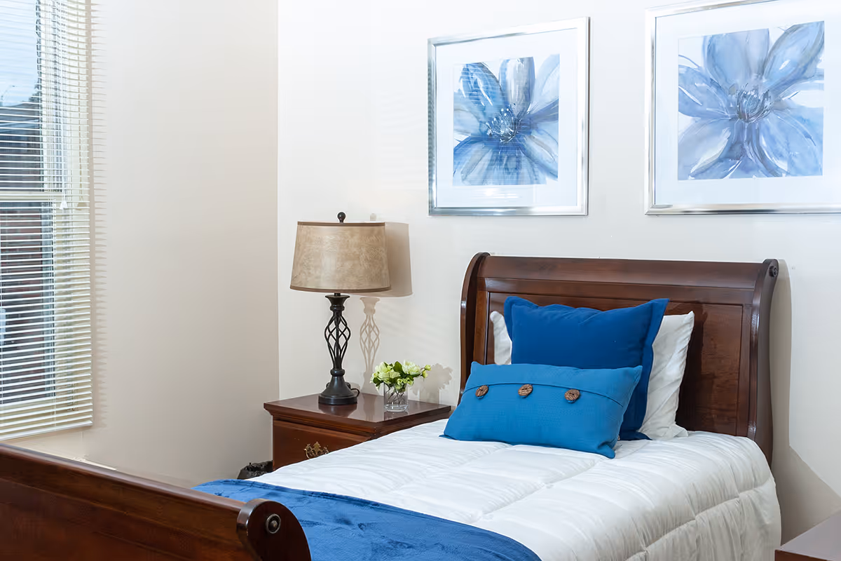 A neatly made single bed with white bedding and two blue pillows in a room. Next to the bed is a wooden nightstand with a decorative lamp and a small vase with flowers. Two framed blue floral paintings hang on the wall above the bed. A window with blinds is visible on the left side.