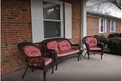 Wicker patio furniture with red patterned cushions on a covered concrete porch outside a brick building.