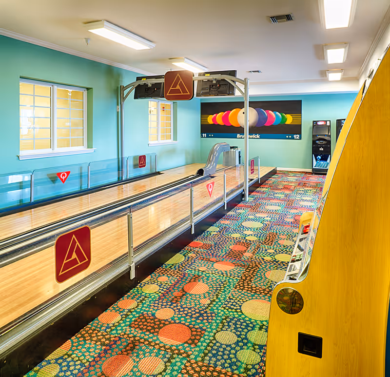Indoor bowling alley with two lanes, colorful patterned carpet, teal walls, and a large painting of bowling balls on the far wall. There are windows on the left wall and arcade machines on the right side near the back.