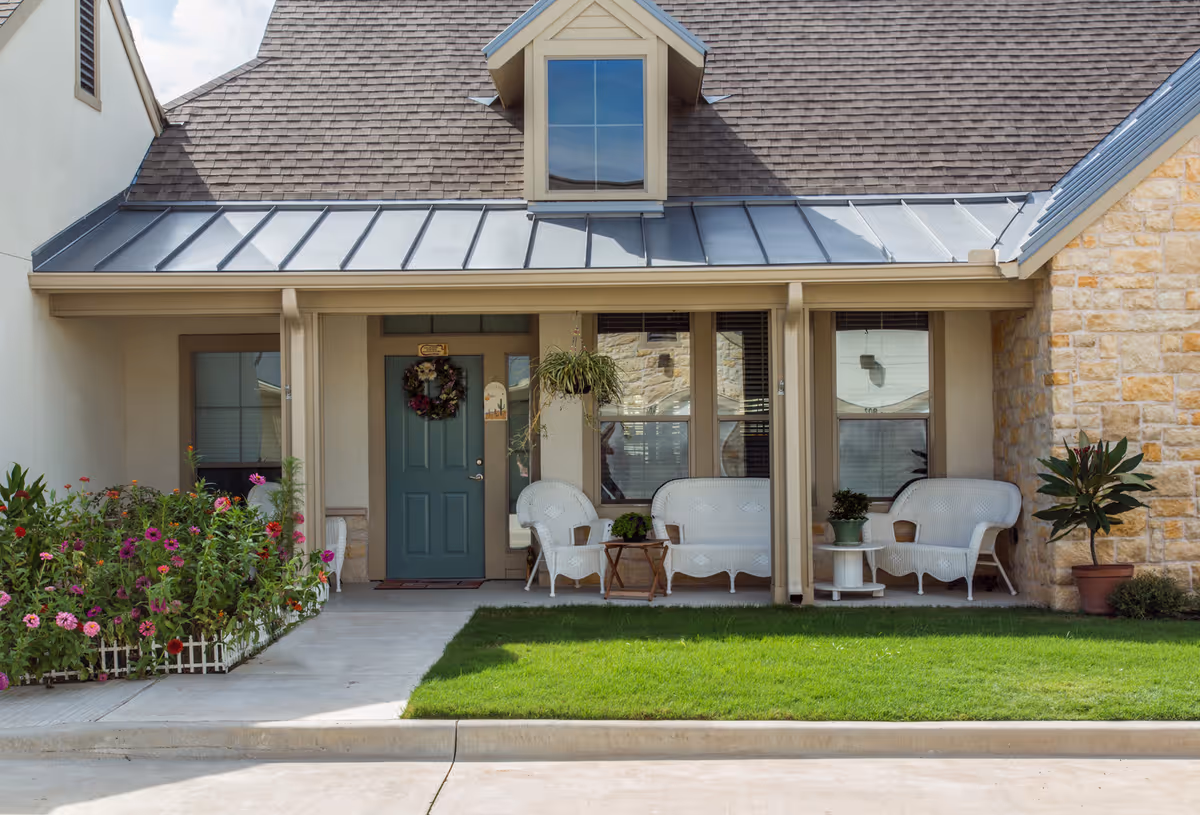 Front porch of a residential building with a green door decorated with a wreath, white wicker chairs and tables, potted plants, a flower bed with colorful flowers, and a well-maintained lawn.