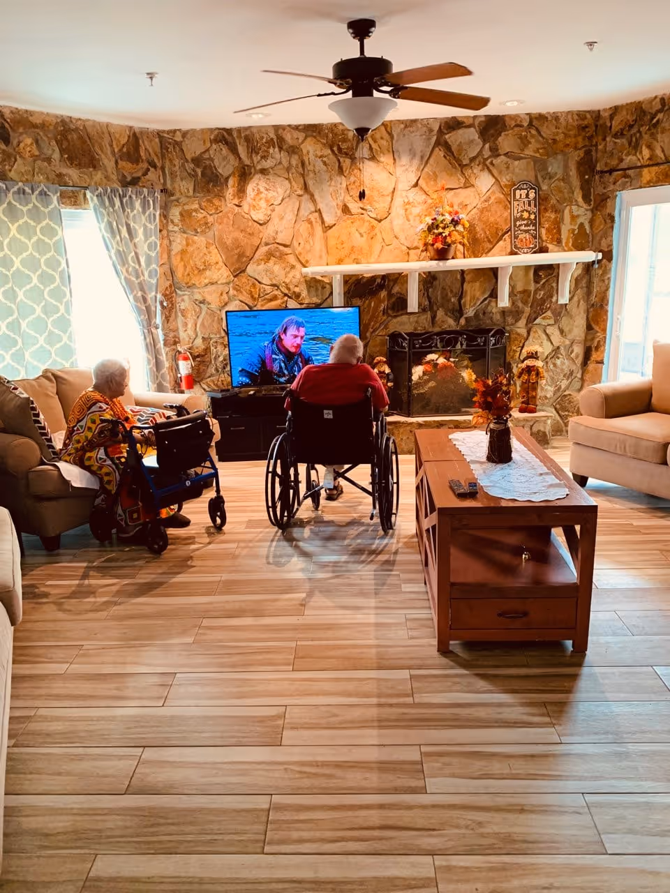 A living room with two elderly residents—one in a wheelchair watching TV and another seated with a walker—facing a stone fireplace, coffee table, and sofas.