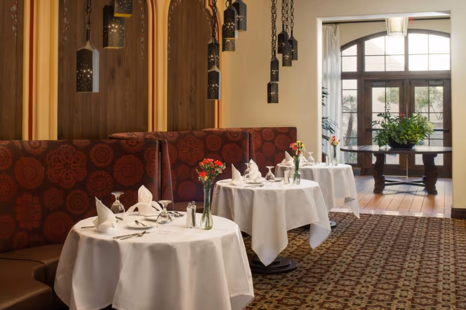 Elegant dining area with round tables covered in white tablecloths, set with folded napkins, glassware, and silverware. Red patterned booth seating lines the wall, and decorative hanging pendant lights illuminate the space. In the background, large glass doors let in natural light and a table with a large green plant is visible.