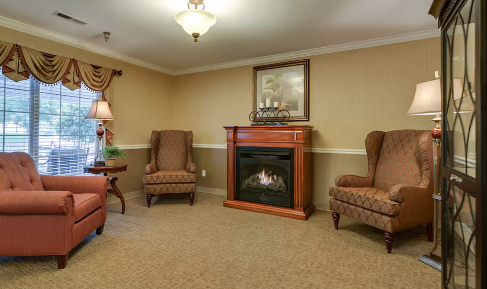 A cozy living room with a gas fireplace in the center, flanked by two patterned armchairs. To the left is a red sofa and a wooden side table with a lamp and a small plant. A large window with decorative curtains lets in natural light. The walls are beige with a white chair rail, and the floor is carpeted.