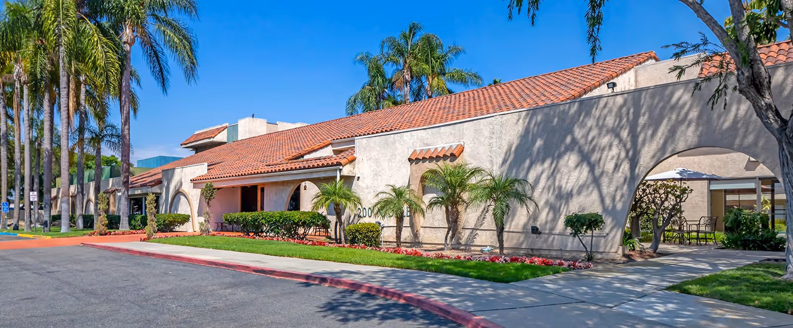 Exterior view of a single-story building with a red tile roof and beige stucco walls, surrounded by palm trees and landscaped greenery under a clear blue sky. There is a sidewalk and a driveway in front of the building.
