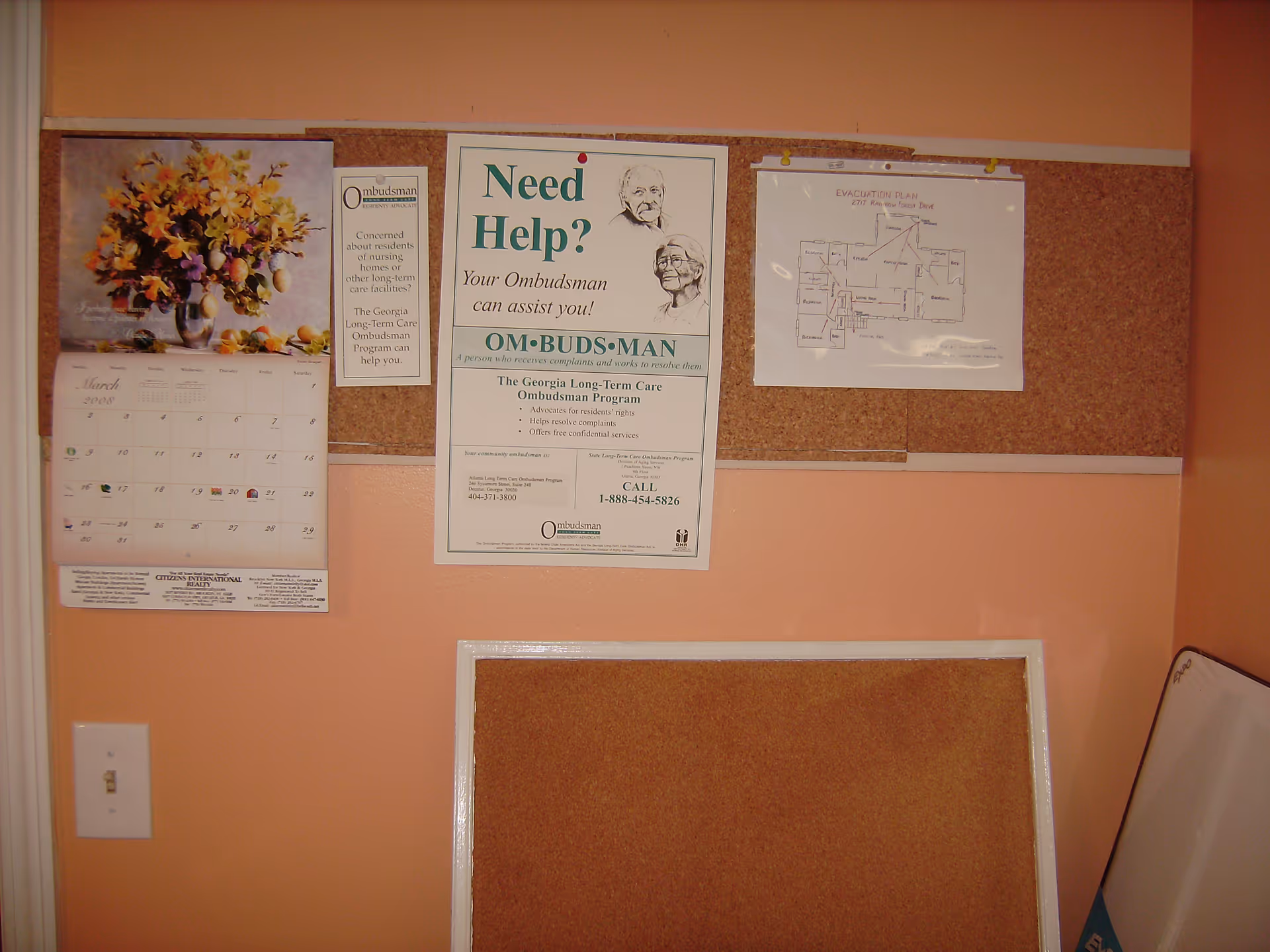 A bulletin board on a peach-colored wall displaying a calendar with a floral image, an informational poster about the Georgia Long-Term Care Ombudsman Program, and an evacuation plan. There is also a light switch on the wall and a smaller empty corkboard below the main bulletin board.
