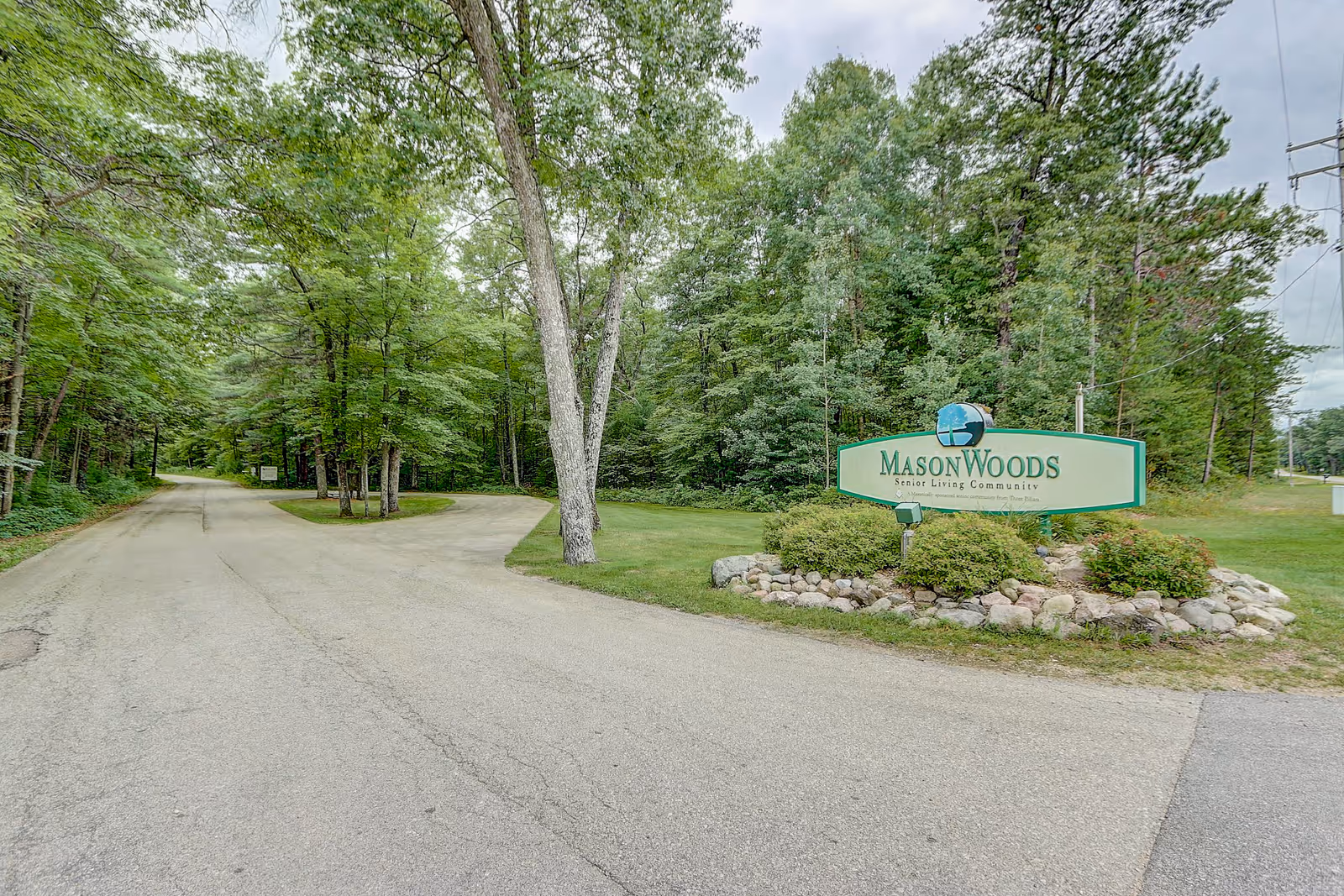 Entrance road to MasonWoods Senior Living Community surrounded by trees and greenery, with a large sign displaying the facility name on the right side of the road.