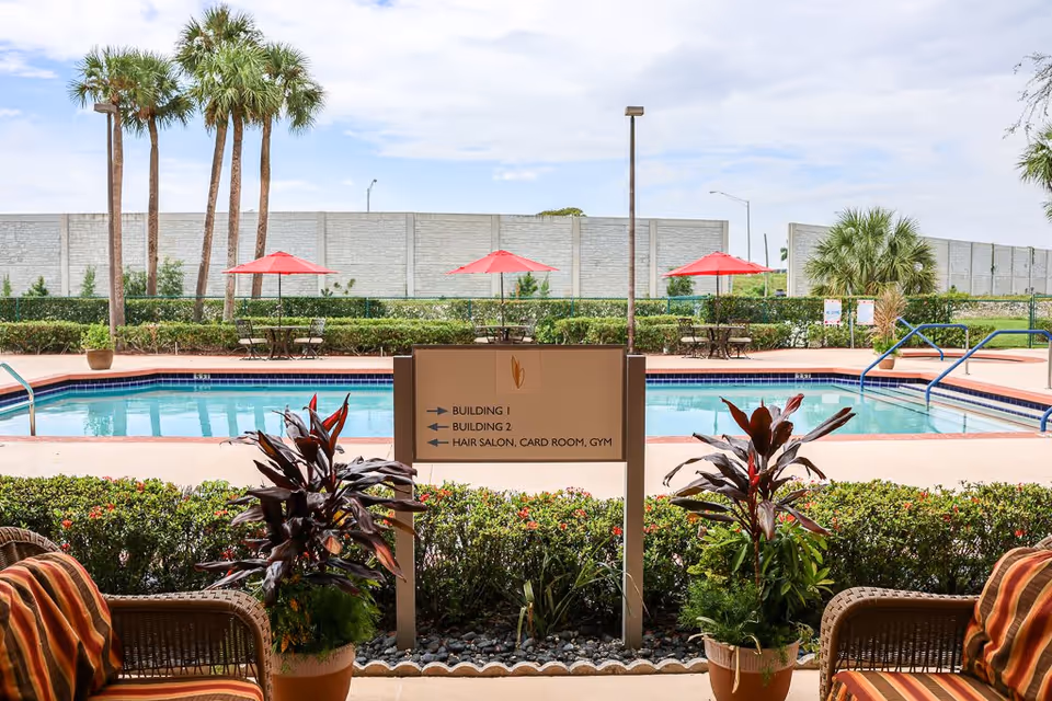 Outdoor view of a swimming pool area with red umbrellas and tables, palm trees, and a directional sign indicating Building 1, Building 2, Hair Salon, Card Room, and Gym. There are two potted plants and wicker chairs with striped cushions in the foreground.