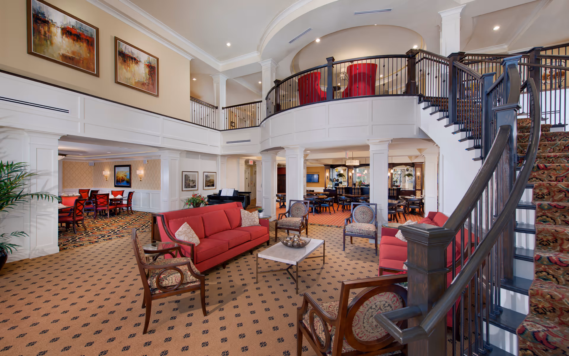 Spacious senior living facility common area with red sofas, patterned armchairs, a marble coffee table, and a grand staircase with dark wood railing and patterned carpet. The room features high ceilings with white columns, a mezzanine with red chairs, and framed artwork on the walls. There is a dining area visible in the background with tables and chairs.